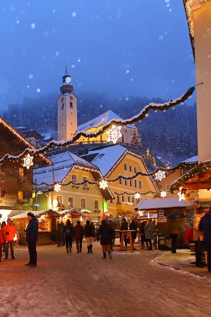 Weihnachtsmarkt mit beleuchteten Holzständen, schneebedecktem Boden und Kirche im Hintergrund bei Abenddämmerung.