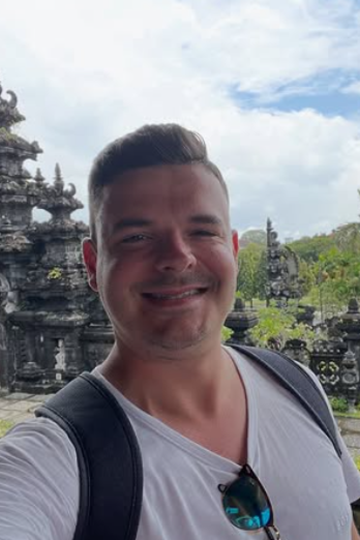 Ein Mann mit Rucksack lächelt vor einem kunstvoll verzierten Tempel mit tropischer Vegetation im Hintergrund.