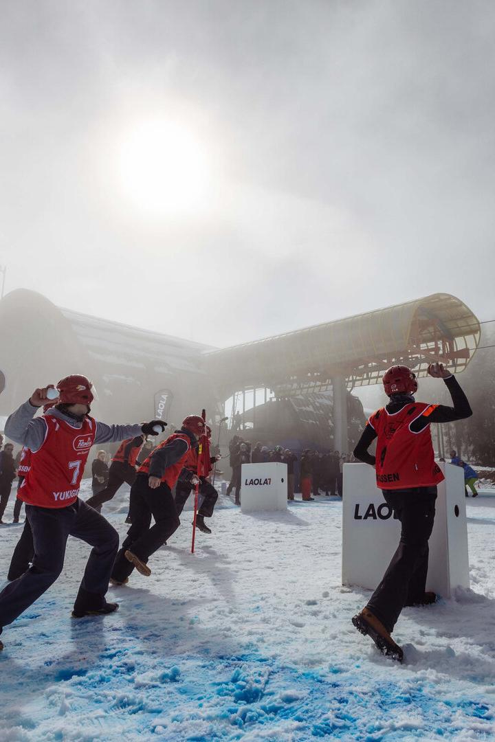 Mehrere Personen in roter Skibekleidung und Helmen stehen auf schneebedecktem Boden vor einer großen, weißen Schneekanone unter bewölktem Himmel.
