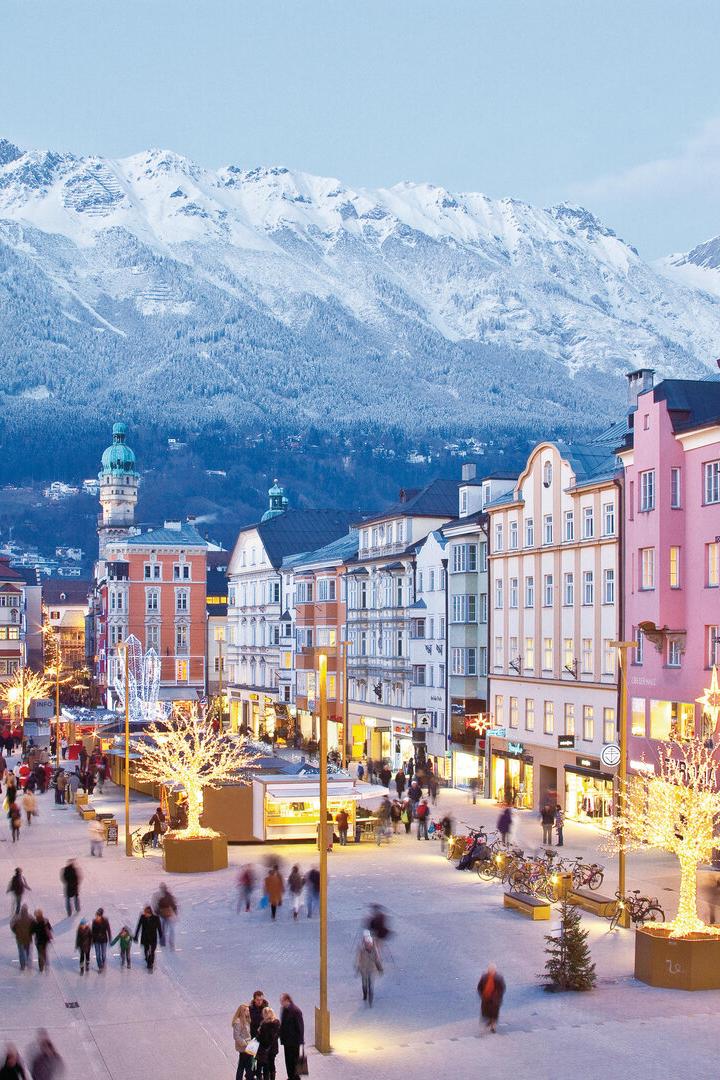 Belebte Fußgängerzone mit beleuchteten Bäumen und historischen Gebäuden vor schneebedeckten Bergen im Abendlicht.