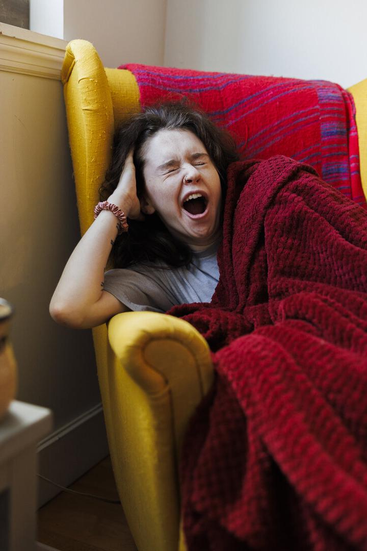 Eine Frau gähnt müde in einem gelben Sessel unter einer roten Decke.