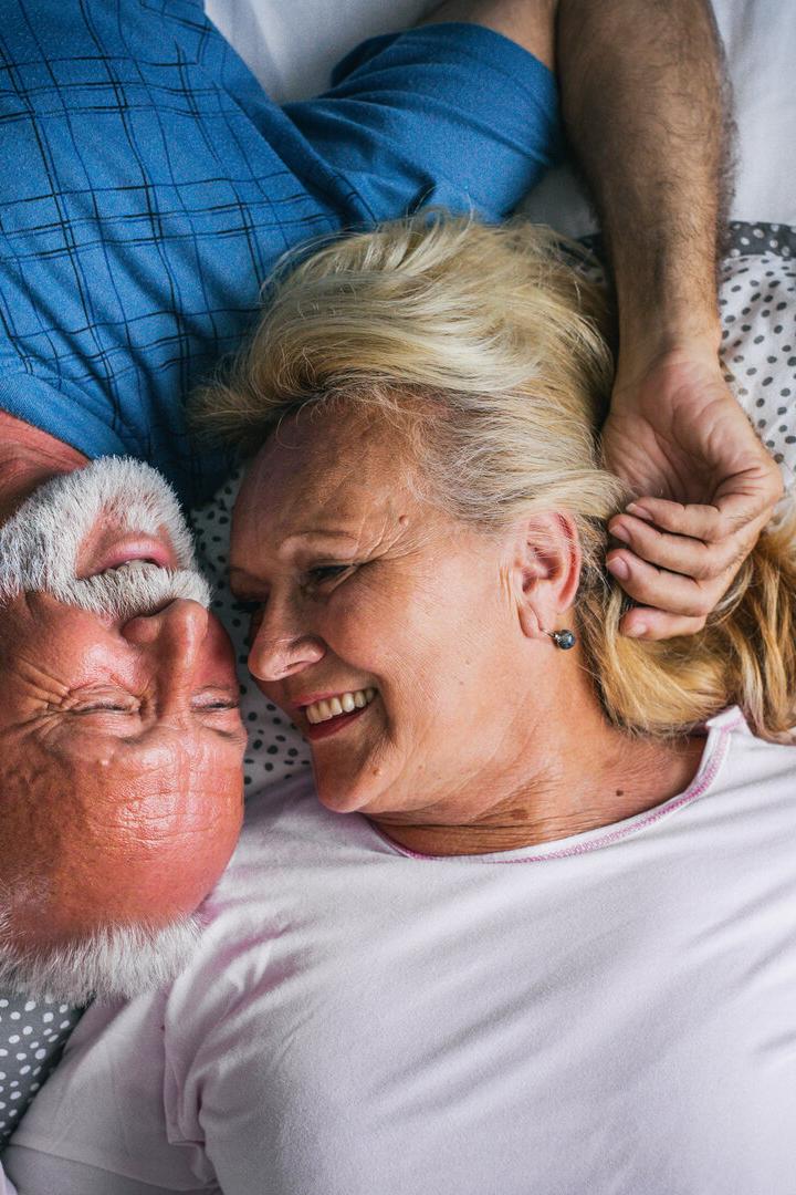 Ein älteres Paar liegt lächelnd im Bett, ihre Gesichter einander zugewandt.