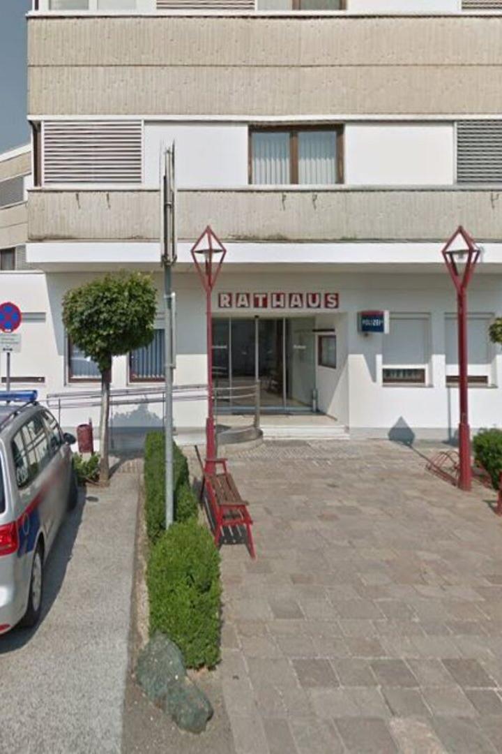 Vor dem Eingang eines Rathauses stehen zwei Polizeiautos, Bänke und kleine Bäume säumen den Weg.