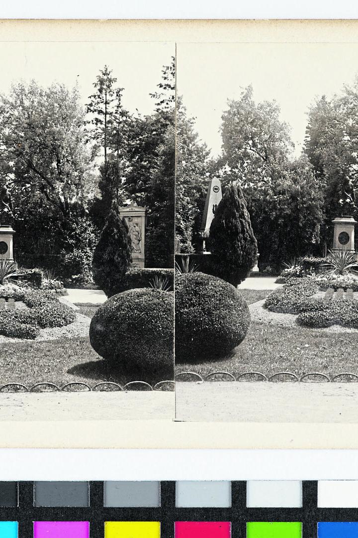 Symmetrisch angelegter Garten mit runden Sträuchern, Blumenbeeten, Denkmälern und Bäumen im Hintergrund.