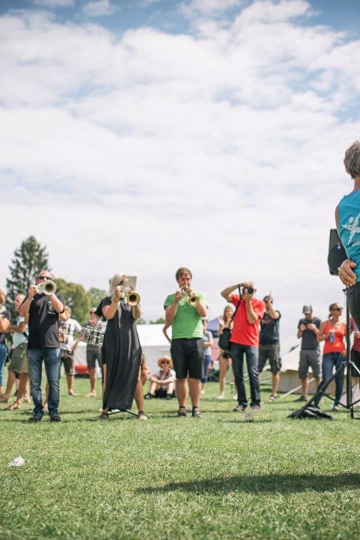 Ein Paar tanzt barfuß auf einer Wiese, während eine Blaskapelle im Hintergrund spielt und Menschen zuschauen.