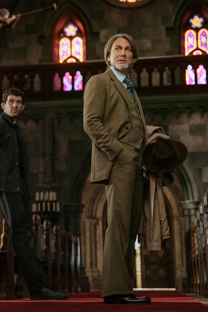 Zwei Männer stehen zwischen Holzbänken in einer Kirche mit bunten Fenstern und schauen in verschiedene Richtungen.