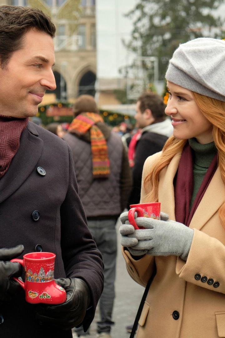 Ein Mann und eine Frau unterhalten sich lächelnd auf einem Weihnachtsmarkt und halten rote Tassen in den Händen.