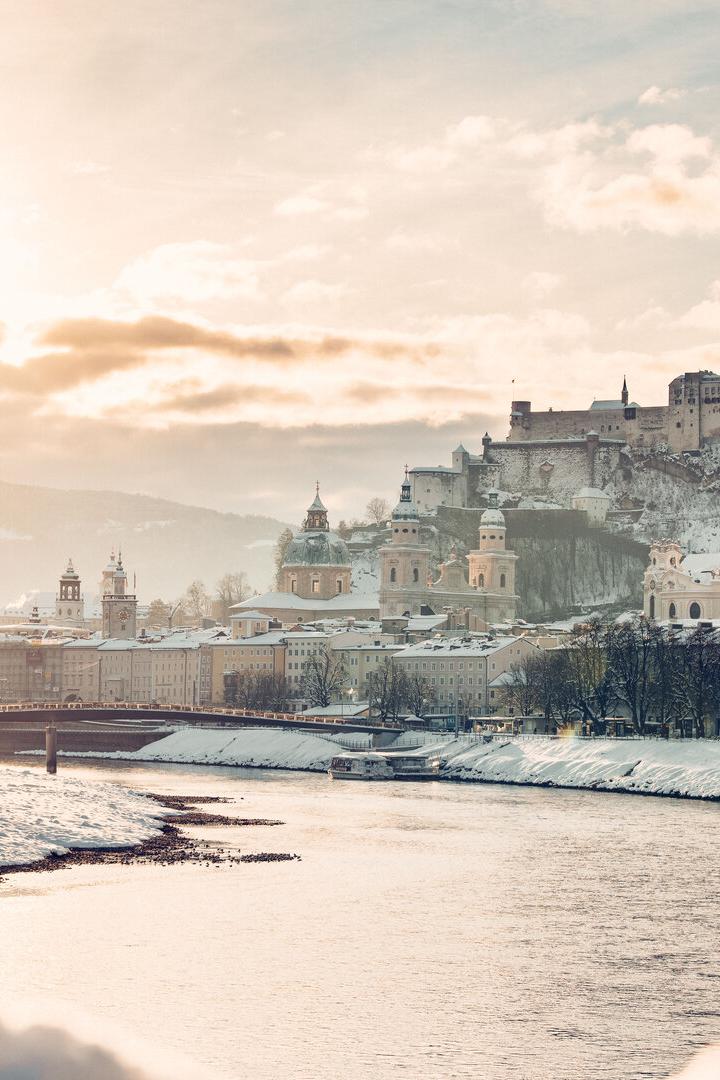 Verschneite Stadt mit Fluss, historischen Gebäuden und einer Burg auf einem Hügel im Sonnenlicht.