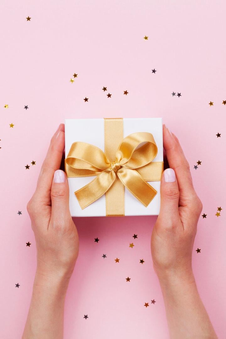 Zwei Hände halten eine weiße Geschenkbox mit goldener Schleife auf rosa Hintergrund mit kleinen Sternkonfetti.