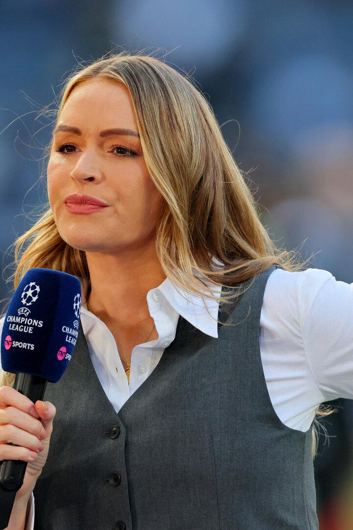 Eine Frau mit langen blonden Haaren hält ein Mikrofon mit Champions-League-Logo und spricht auf einem Sportplatz.