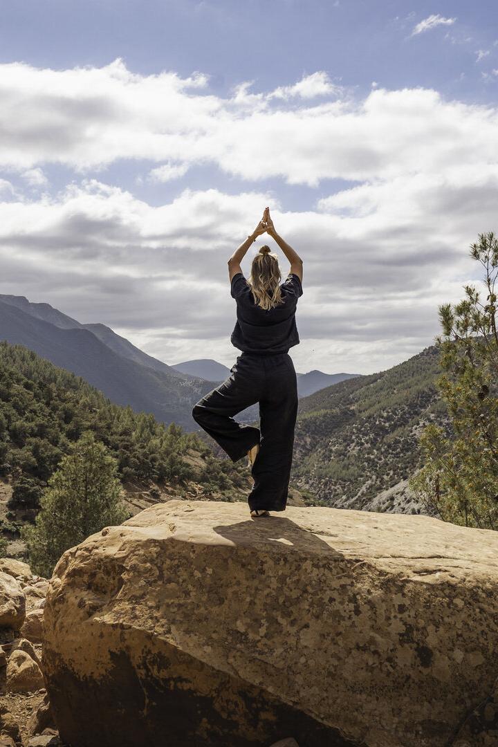One woman practising yoga in tree pose on rock overlooking forested mountains in nature in the Atlas Mountains, Ourika Valley, Morocco