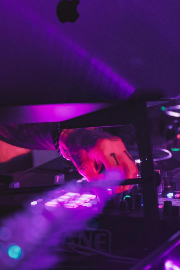 Ein DJ mit Tätowierung bedient ein Pioneer-Mischpult unter violettem Licht, ein Laptop steht daneben.