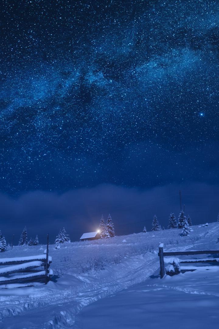Verschneite Landschaft mit Tannen, einer Hütte mit Licht und einem klaren Sternenhimmel in der Nacht.