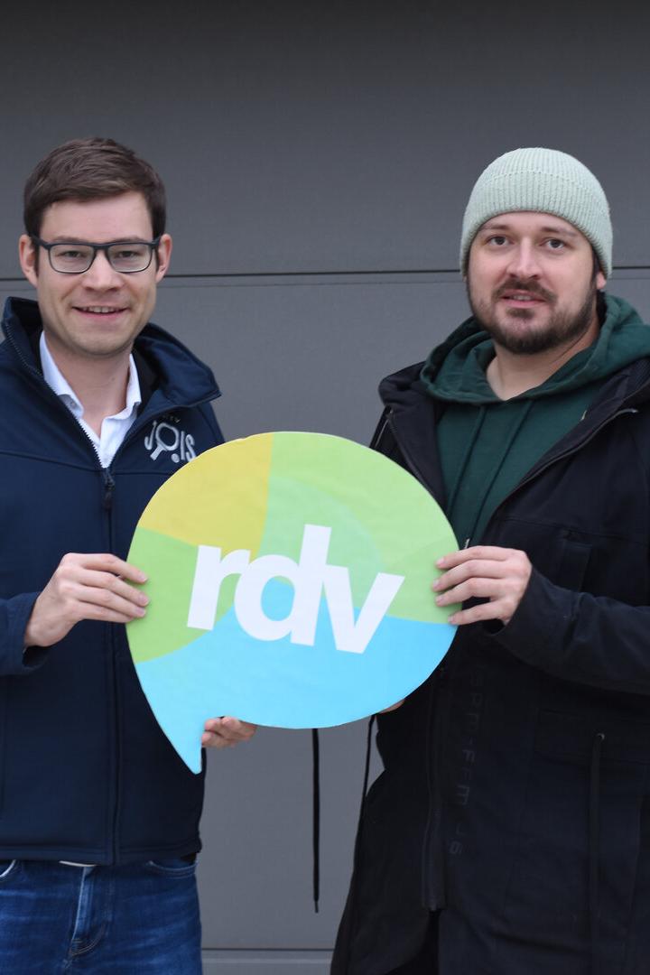 Zwei Männer stehen nebeneinander und halten gemeinsam ein rundes Schild mit der Aufschrift „rdv“.