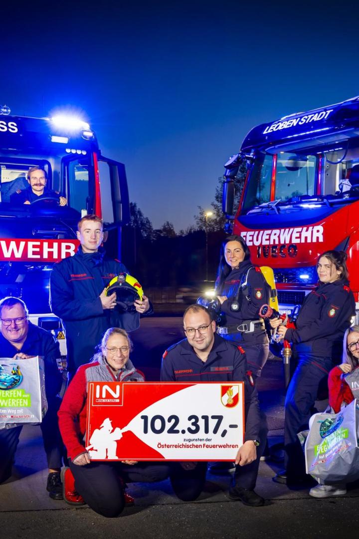 Feuerwehrleute und Interspar-Mitarbeitende präsentieren vor zwei Feuerwehrautos einen Scheck über 102.317 Euro, gespendet durch Pfandflaschenaktionen.