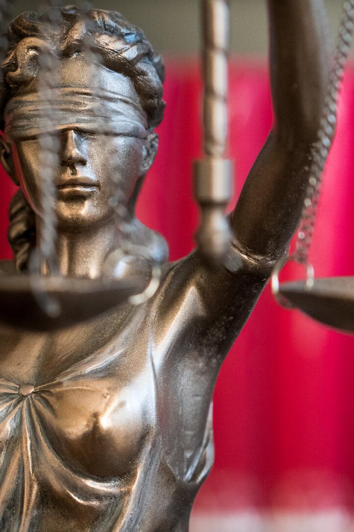 Eine Justitia-Statue mit Augenbinde und Waage steht vor einem roten Hintergrund.
