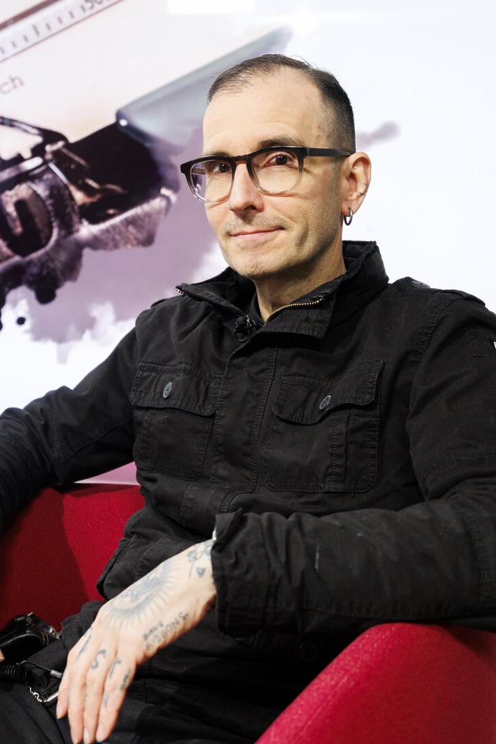 Ein Mann mit Brille und Tattoos sitzt in einem roten Sessel, im Hintergrund eine Schreibmaschine und eine pinke Orchidee.