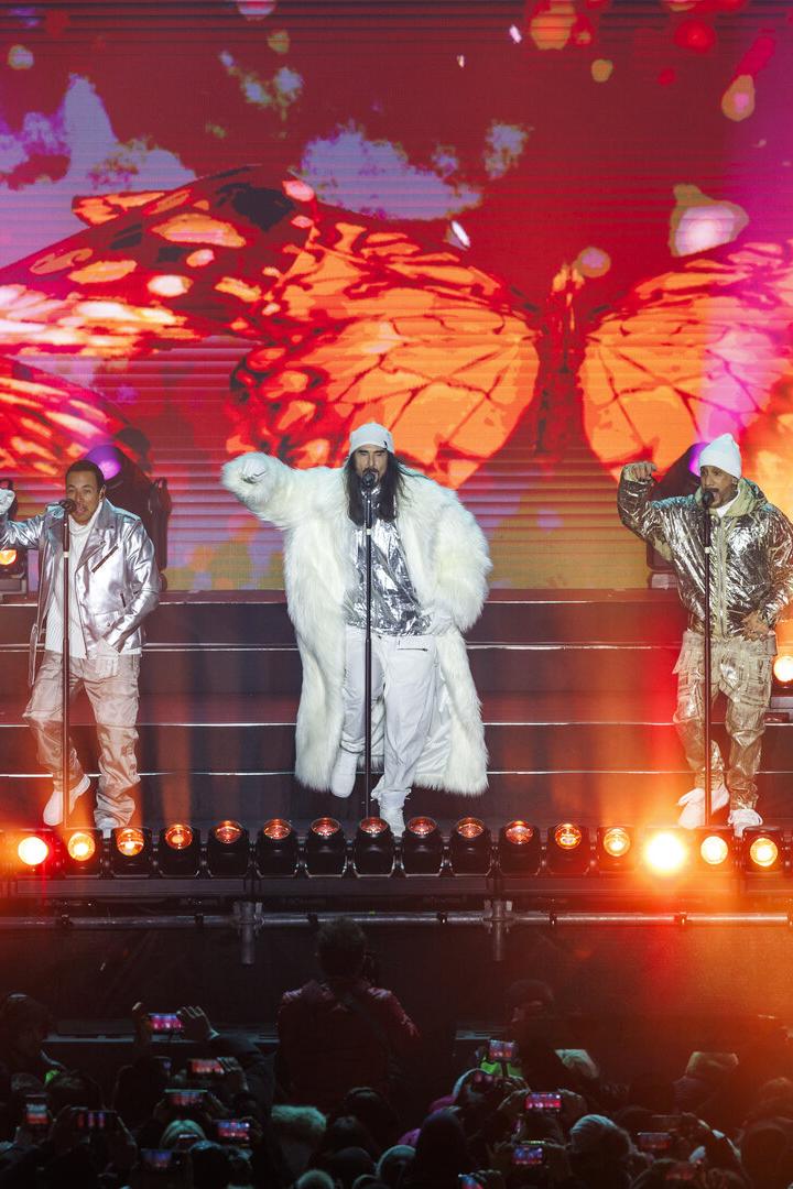 Fünf Sänger in silbernen Outfits stehen auf einer Bühne mit bunten Lichtern und Schmetterlingsprojektion im Hintergrund.