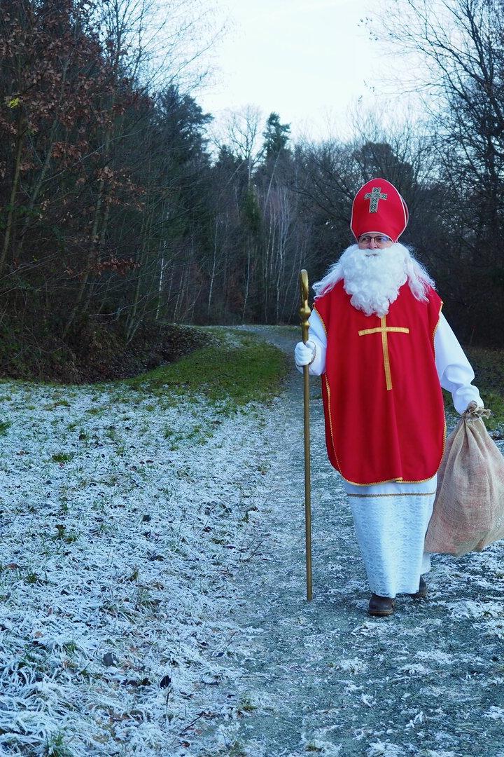 Eine als Nikolaus verkleidete Person mit rotem Mantel, Bischofsmütze und Stab geht auf einem frostigen Waldweg.
