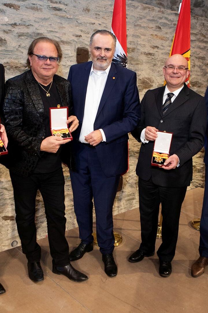 Fünf Männer in Anzügen stehen nebeneinander und halten Auszeichnungen vor sich, im Hintergrund sind Fahnen zu sehen.