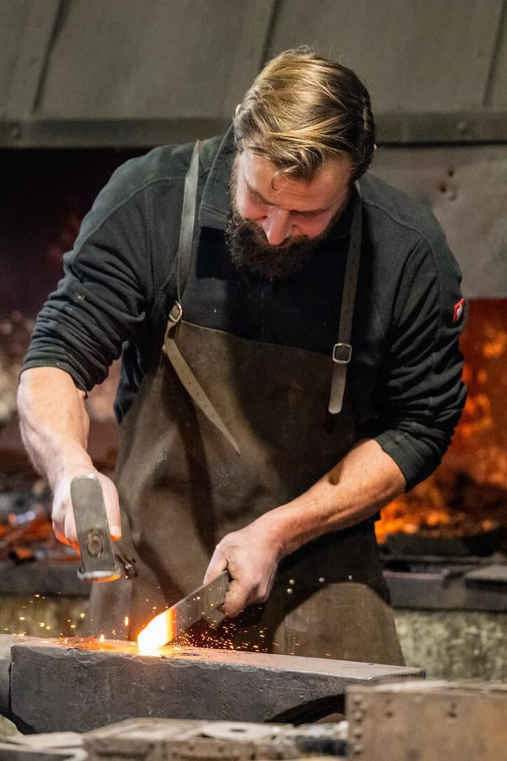 Ein Schmied mit Bart bearbeitet glühendes Metall auf einem Amboss mit Hammer.