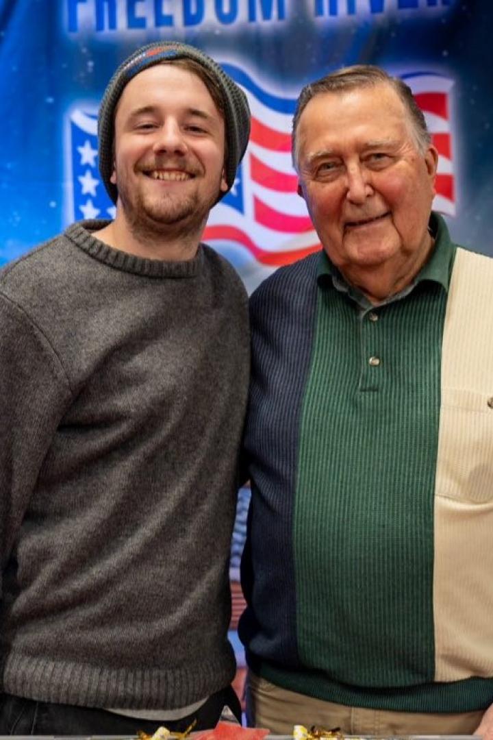 Zwei Männer lächeln vor einer festlichen Ballon-Dekoration mit US-Flagge im Hintergrund.