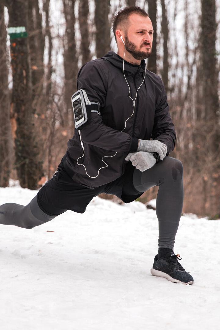 Ein Mann macht einen Ausfallschritt im verschneiten Wald und trägt Sportkleidung sowie Kopfhörer.