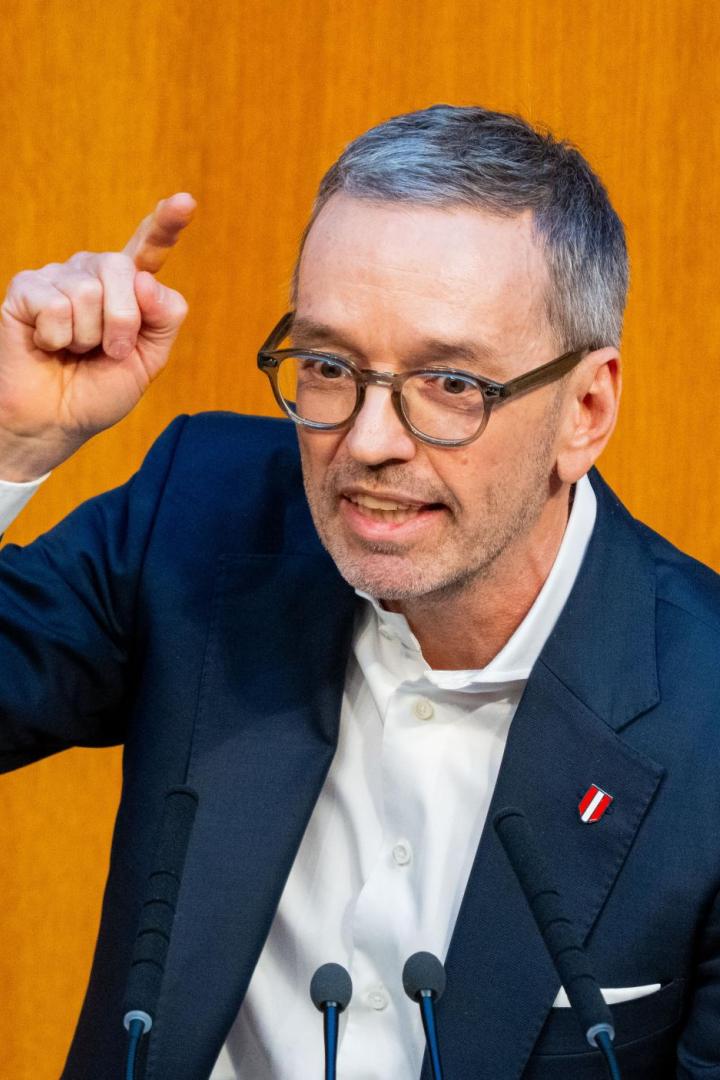 Ein Mann mit Brille gestikuliert vor einem orangefarbenen Hintergrund mit Mikrofonen.