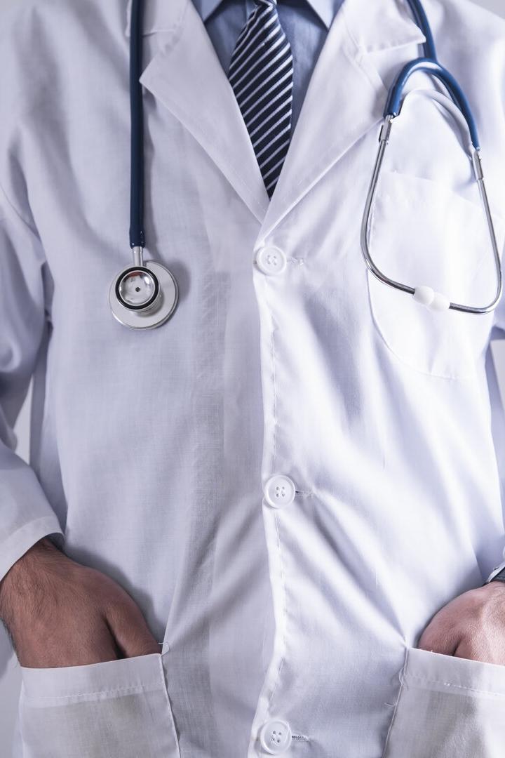 Ein Arzt im weißen Kittel mit Stethoskop und gestreifter Krawatte steht mit den Händen in den Taschen da.