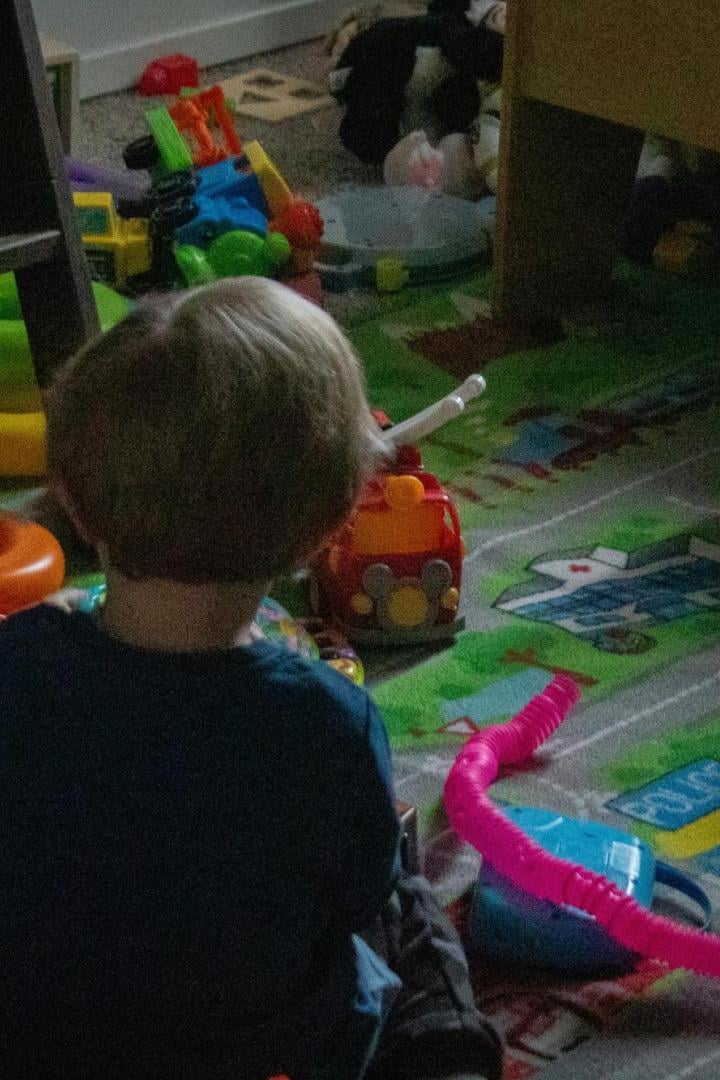 Ein kleines Kind sitzt in einem unaufgeräumten Kinderzimmer voller Spielzeug auf dem Boden.
