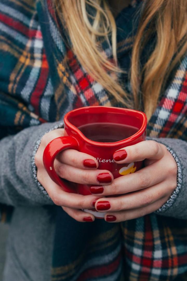 Eine Person mit rot lackierten Nägeln hält eine rote Tasse mit Heißgetränk und trägt einen karierten Schal.