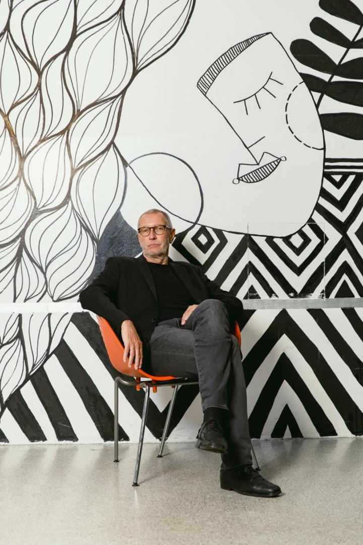 Konrad Paul Liessmann sitzt entspannt auf einem Stuhl vor einer schwarz-weiß gestalteten Wand mit abstrakten Mustern und Zeichnungen.