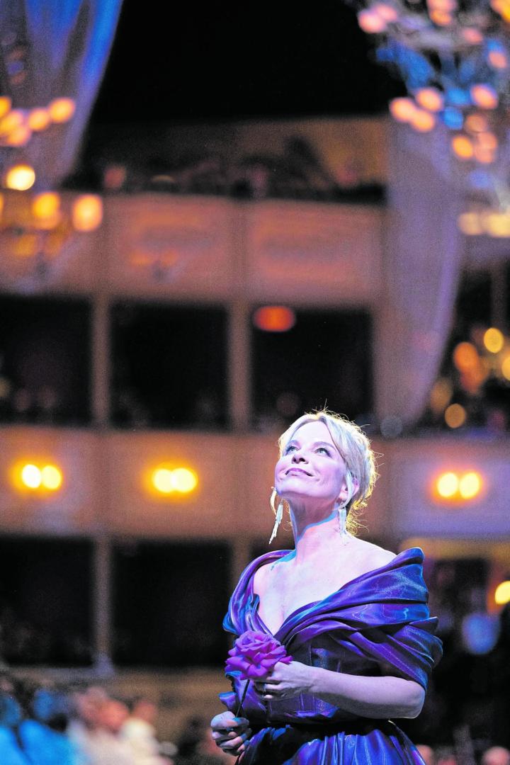 Eine elegant gekleidete Frau in einem lila Kleid hält eine Blume und blickt lächelnd nach oben in einem festlich beleuchteten Saal.