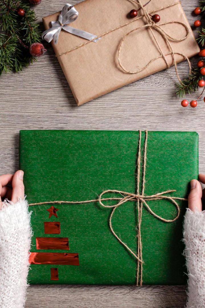 Verschieden verpackte Weihnachtsgeschenke mit Naturdeko und Tannenzweigen liegen auf einem Holztisch, zwei Hände halten ein grün verpacktes Geschenk mit Schleife.