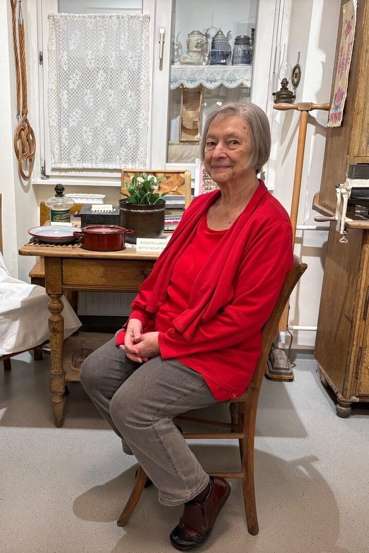 Eine ältere Frau mit grauen Haaren und rotem Oberteil sitzt in einer gemütlichen, altmodischen Küche auf einem Stuhl.