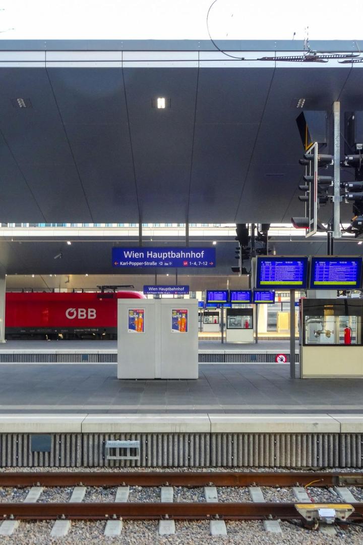 Bahnsteig am Wiener Hauptbahnhof mit Anzeigetafeln, rotem ÖBB-Zug und moderner Überdachung.
