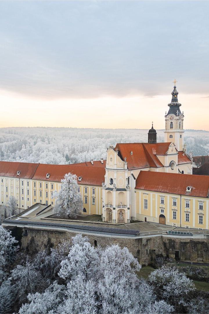 Das Stift Altenburg mit rotem Dach und Kirchturm liegt inmitten einer winterlichen, schneebedeckten Landschaft.