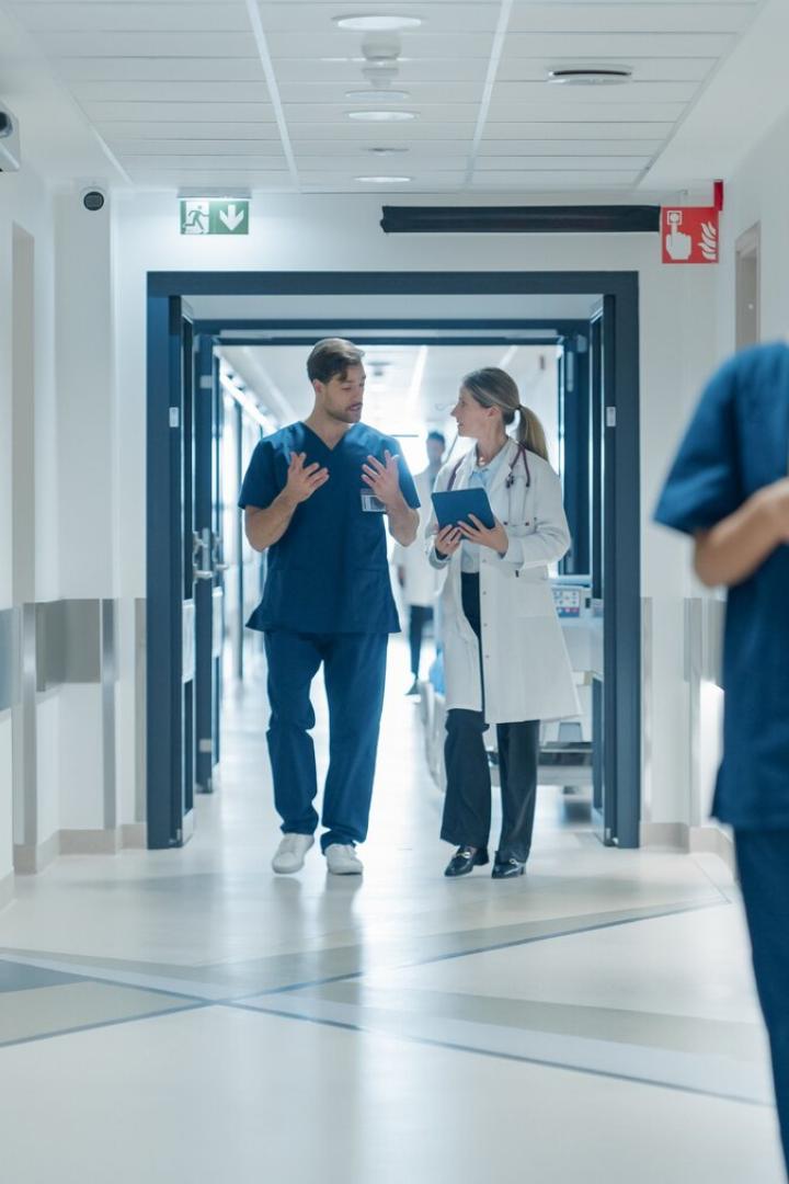 Healthcare Professionals Walking to Their Examination Rooms, Reception Desk, ICU Units in a Modern Bright Hospital Corridor. Female Doctor Showing a Young Male Surgeon Information on a Tablet