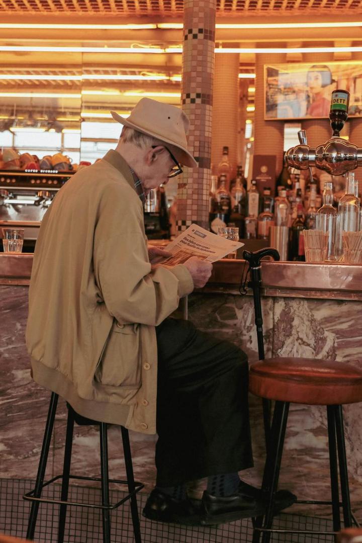 Ein älterer Mann mit Hut sitzt an einer Bar, liest eine Zeitung und hat einen Gehstock neben sich abgestellt.
