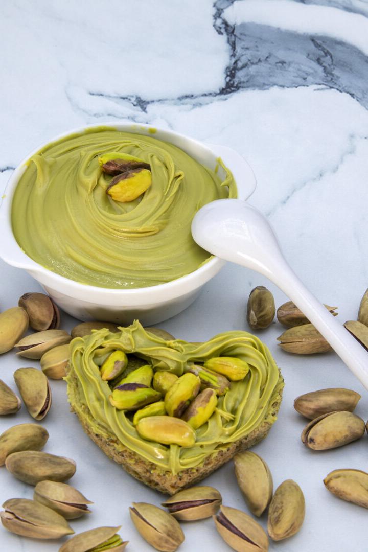 Herzförmiges Brotstück mit Pistaziencreme und Pistazien, daneben eine Schale mit Creme und ein Löffel, umgeben von Pistazien.