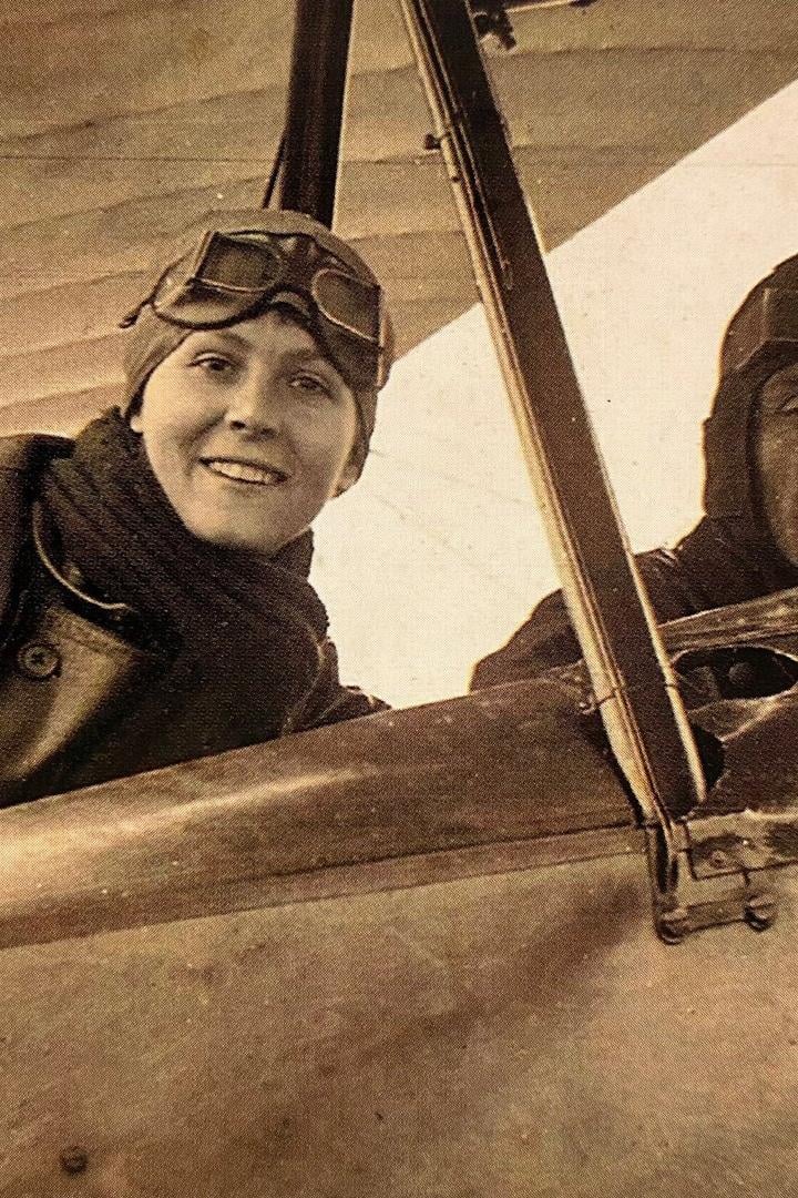 Zwei Personen mit Fliegermützen und Schutzbrillen sitzen in einem offenen Doppeldeckerflugzeug.