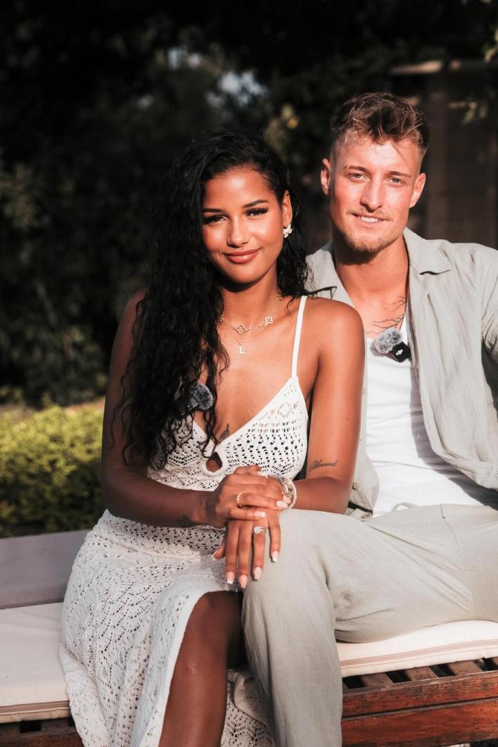 Brenda und Laurenz sitzen eng beieinander auf einer Bank im Freien und lächeln in die Kamera.