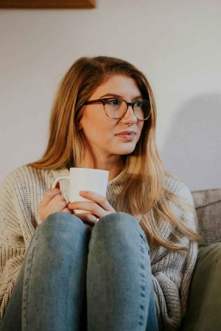 Frau mit Brille sitzt nachdenklich mit einer Tasse auf einem Sofa.