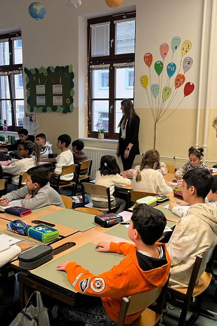 Viele Kinder sitzen an Tischen in einem Klassenraum, während zwei Erwachsene sie betreuen und unterrichten.