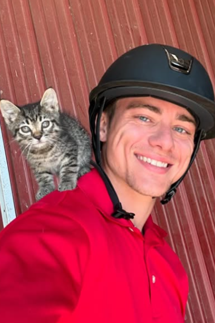 Mann in rotem T-Shirt steht mit einer Katze auf seiner Schulter und einem schwarzen Reithelm auf dem Kopf vor einer Wand