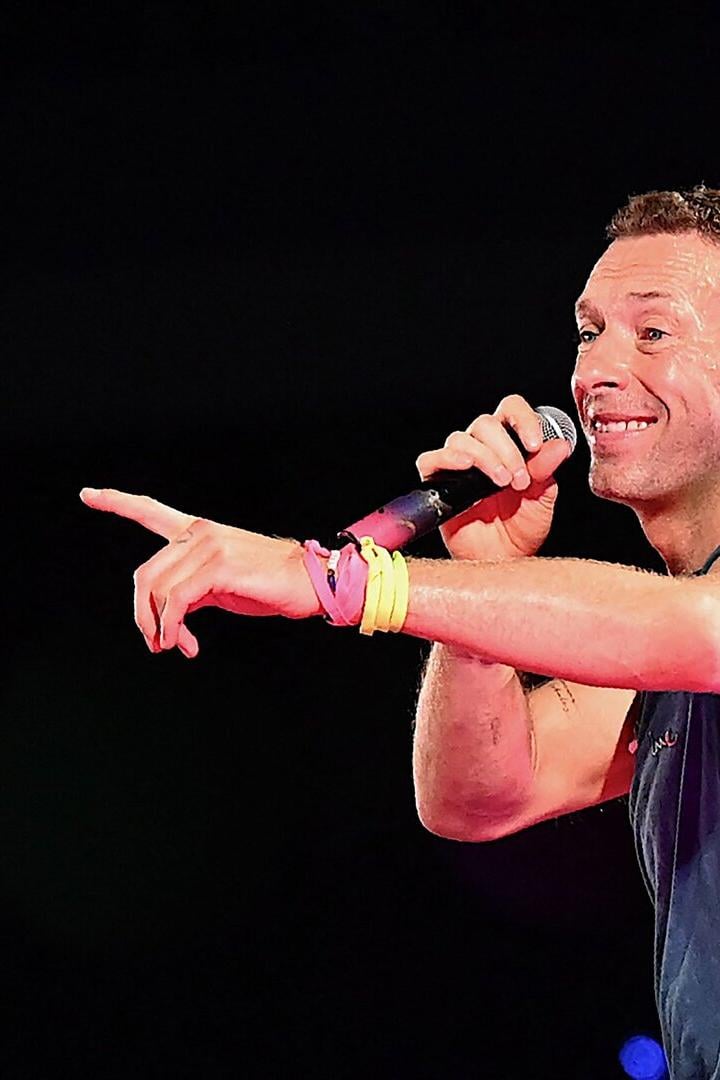 Chris Martin singt mit Mikrofon und zeigt mit dem Finger auf der Bühne.