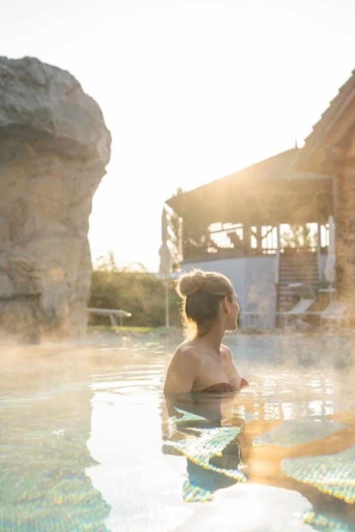 Zwei Personen entspannen im dampfenden Außenpool vor einer Holzhütte bei Sonnenuntergang.