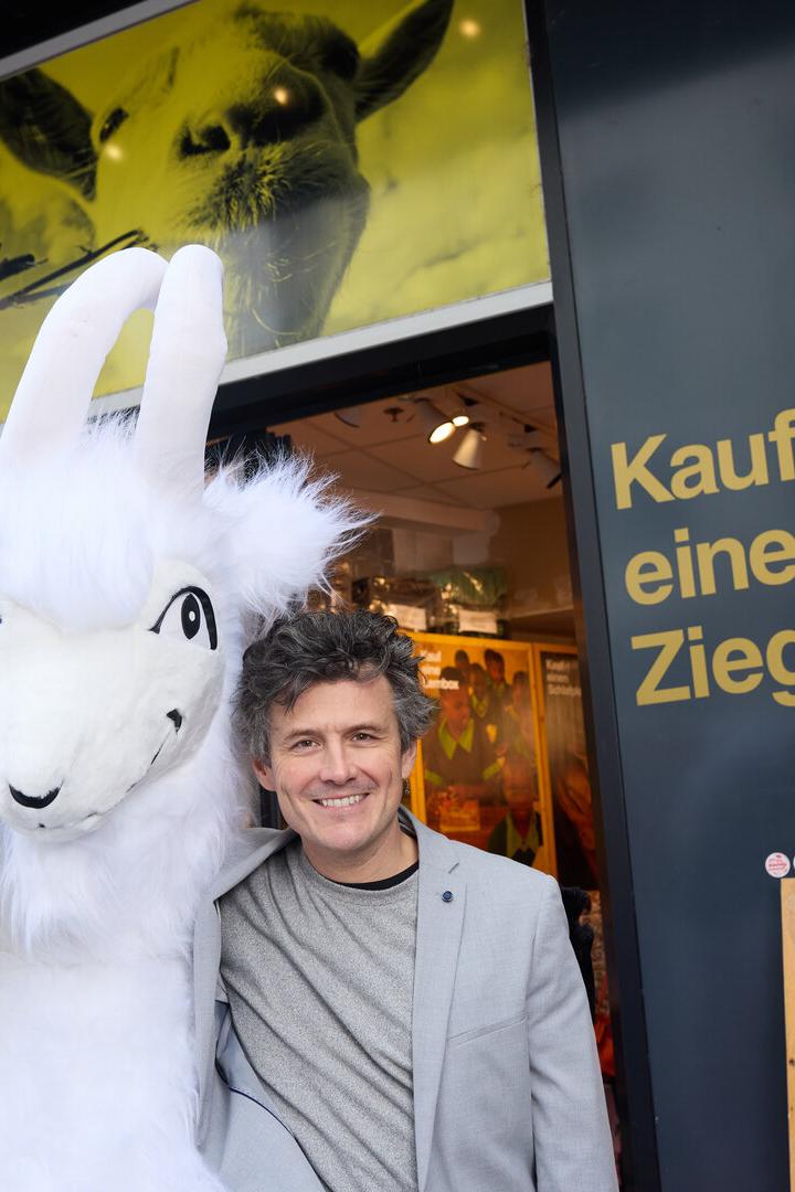 Ein Mann lächelt neben einem Ziegen-Maskottchen vor einem Laden mit der Aufschrift "Kauf eine Ziege".