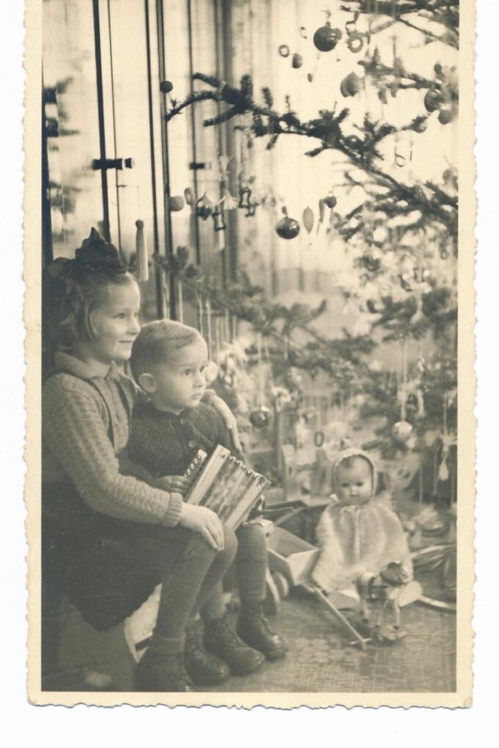 Drei Kinder sitzen neben einem geschmückten Weihnachtsbaum, eines hält ein Akkordeon, im Hintergrund liegt Spielzeug.