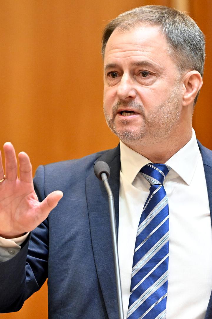 Ein Mann im Anzug spricht am Rednerpult und hebt die Hand vor warmem Holz-Hintergrund.